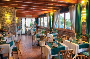 A restaurant with wooden beams, stone walls, and green curtains. Tables are set with white tablecloths and candles. - Hotel Ariston & Palazzo Santa Caterina (Taormina)