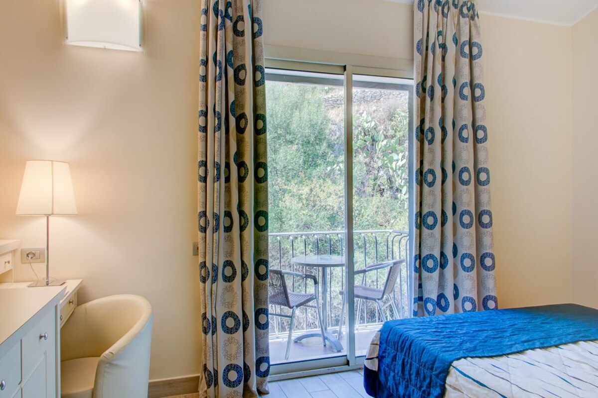 A bedroom with a bed, a desk, a chair, and a balcony with a view of greenery.