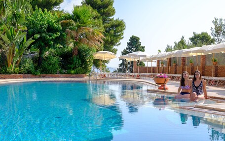 A swimming pool with two people sitting on the edge, surrounded by lush greenery and umbrellas.