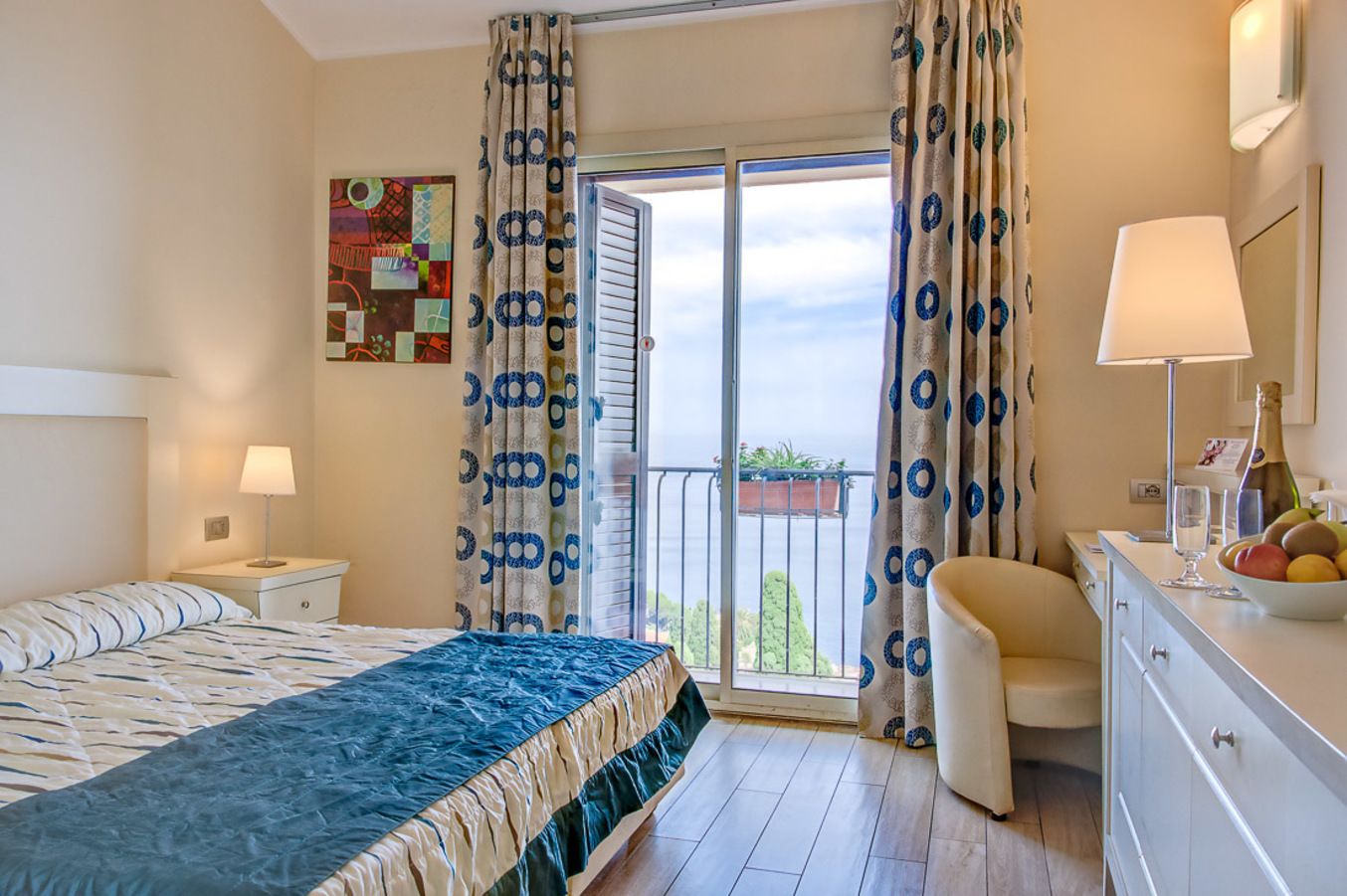 A hotel room with a bed, a desk with a lamp, a chair, a balcony with curtains, and a view of the sea.