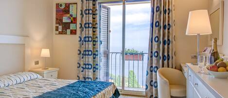 A hotel room with a bed, a desk with a lamp, a chair, a balcony with curtains, and a view of the sea.