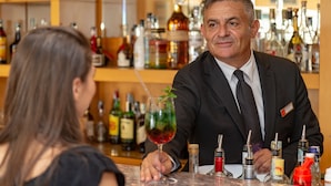 Una zona de bar con un hombre en traje y una mujer sosteniendo una copa. Hay varias botellas y utensilios de bar sobre la barra.