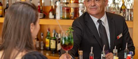 A bar area with a man in a suit and a woman holding a glass. There are various bottles and bar tools on the counter.