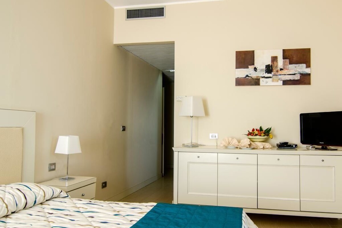 A hotel room with a bed, a bedside table with a lamp, a TV, a cabinet, and artwork on the wall.