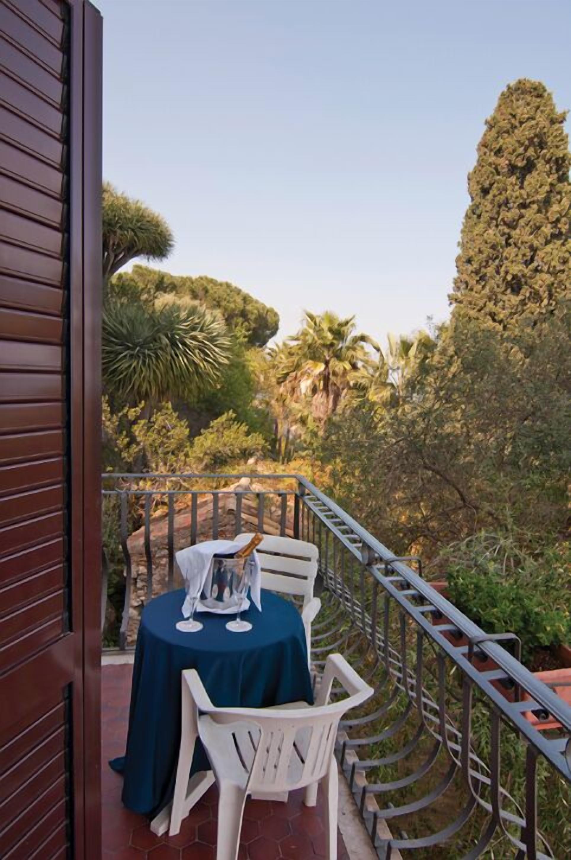 A balcony with a table set for two, surrounded by a metal railing, overlooking a lush garden with various trees and plants.