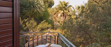 A balcony with a table set for two, surrounded by a metal railing, overlooking a lush garden with various trees and plants.