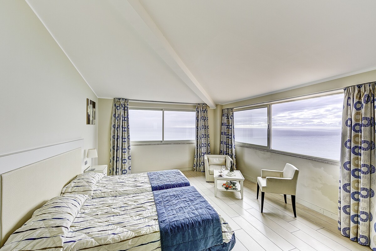 A hotel room with a large bed, a chair, a small table, and a view of the sea through the windows.
