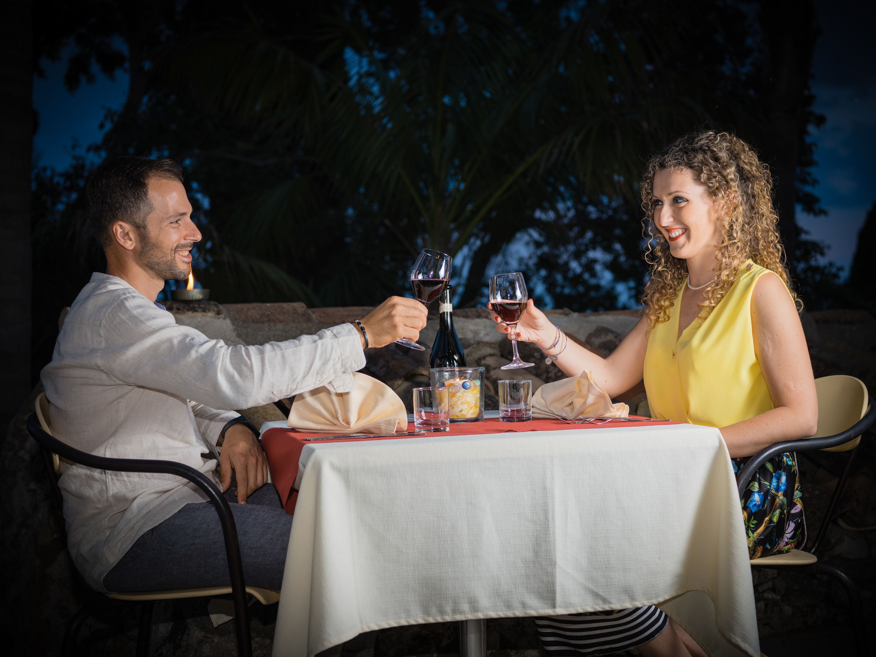 Duas pessoas brindando com taças de vinho em uma mesa ao ar livre com toalha branca, cercadas por cadeiras e uma garrafa de vinho.