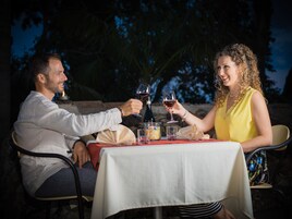 Duas pessoas brindando com taças de vinho em uma mesa ao ar livre com toalha branca, cercadas por cadeiras e uma garrafa de vinho.