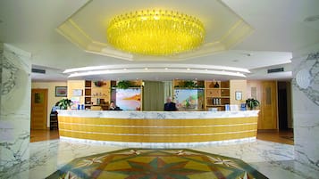 Ein Hotel-Empfangsbereich mit Marmorboden, einer großen Chandelüster und einem Empfangstresen, an dem zwei Mitarbeiter arbeiten.