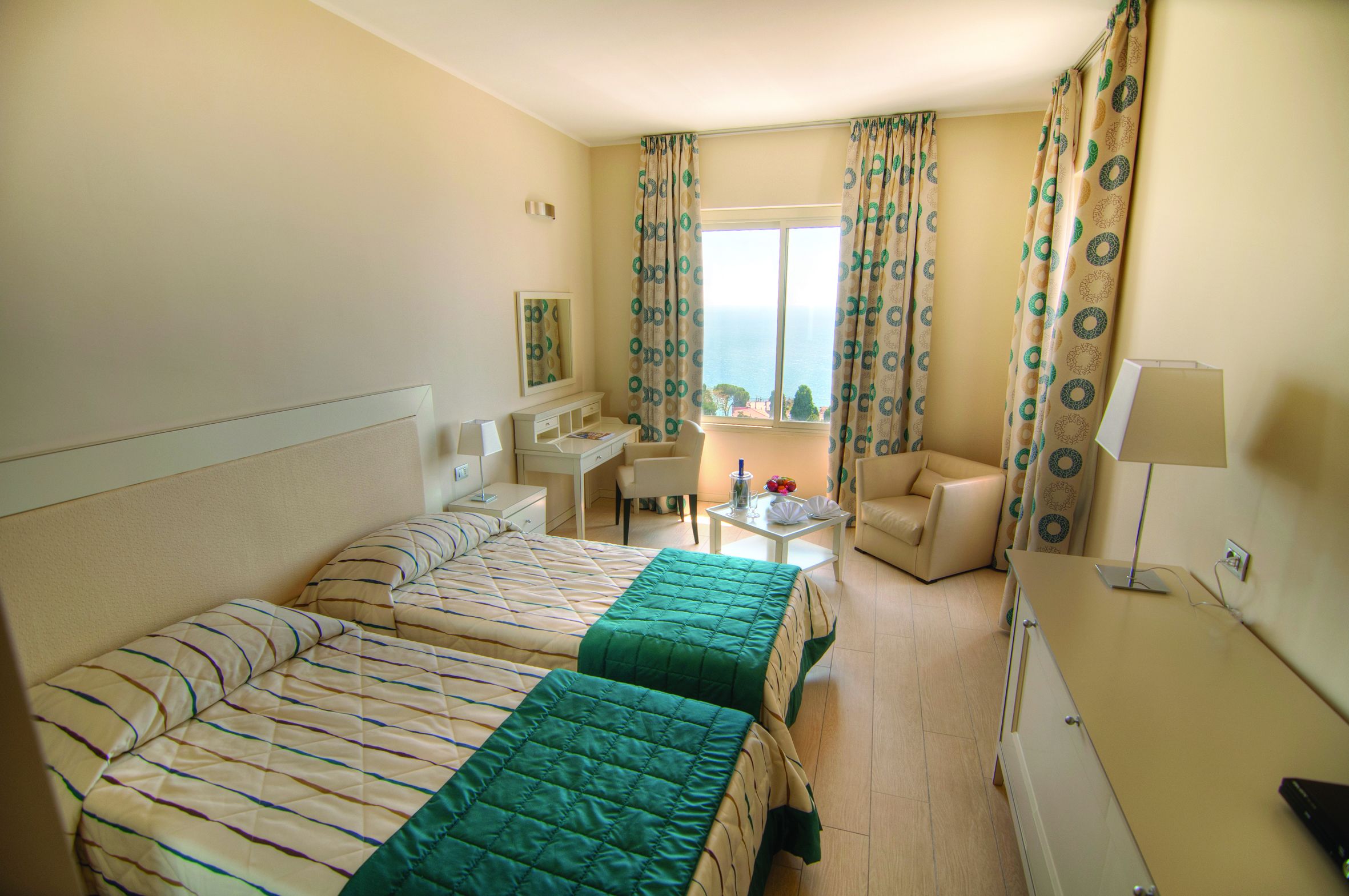 A hotel room with two beds, a desk, a chair, a lamp, and a view of the sea.
