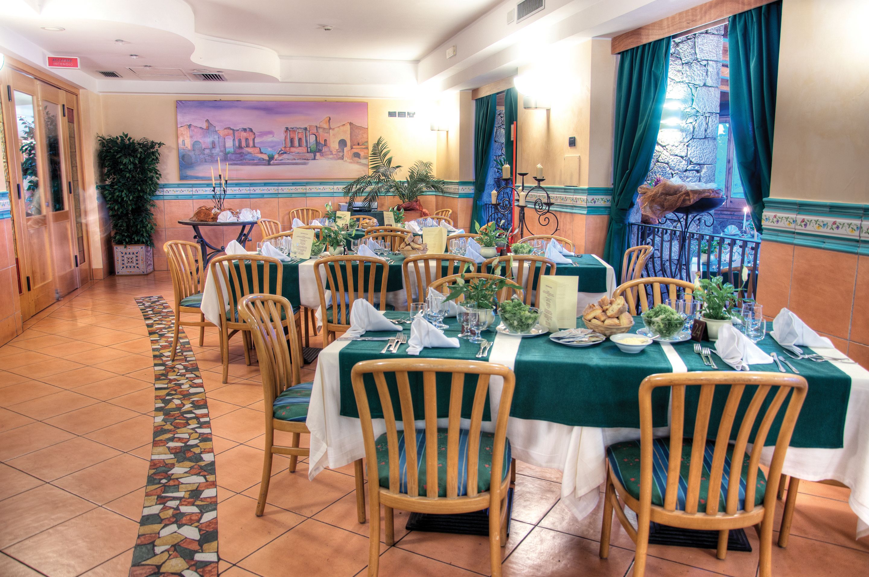 A dining area with multiple tables set for service, chairs, a large painting on the wall, and a view through a window with curtains.