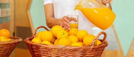 A person pouring orange juice into a glass from a pitcher, with baskets of oranges on a table.