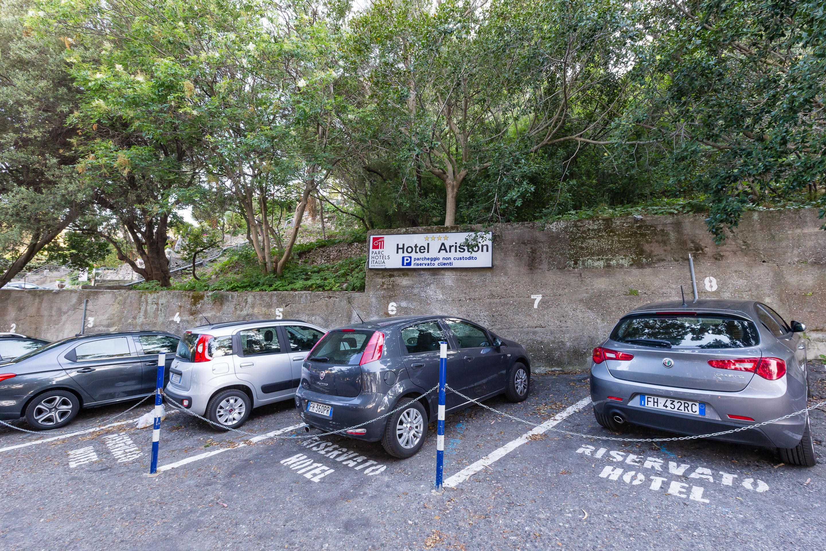 A parking area with a sign for Hotel Ariston, marked parking spaces, and a concrete wall with trees.