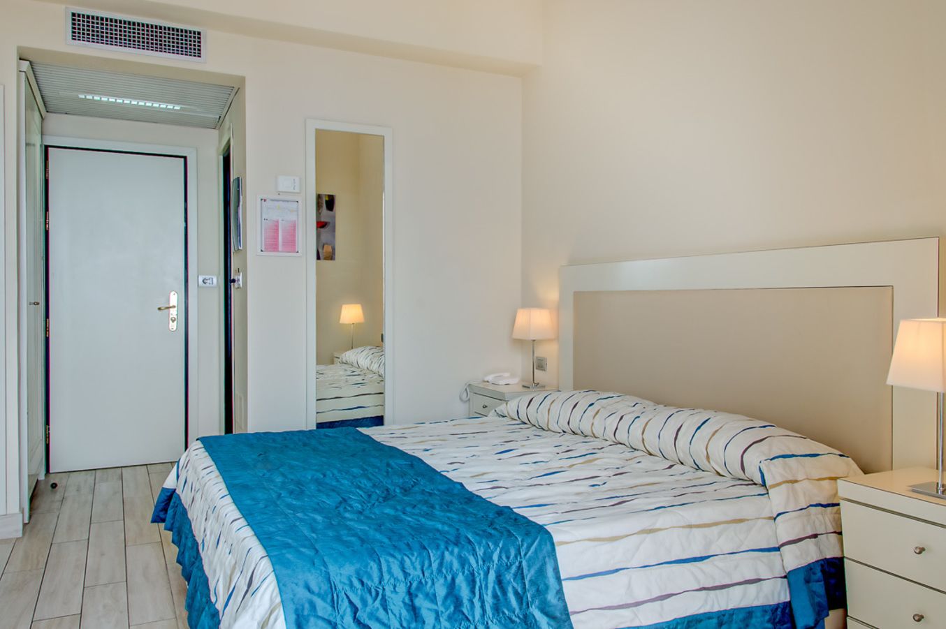 Una habitación de hotel con cama, mesitas de noche, una puerta y una lámpara fijada a la pared.