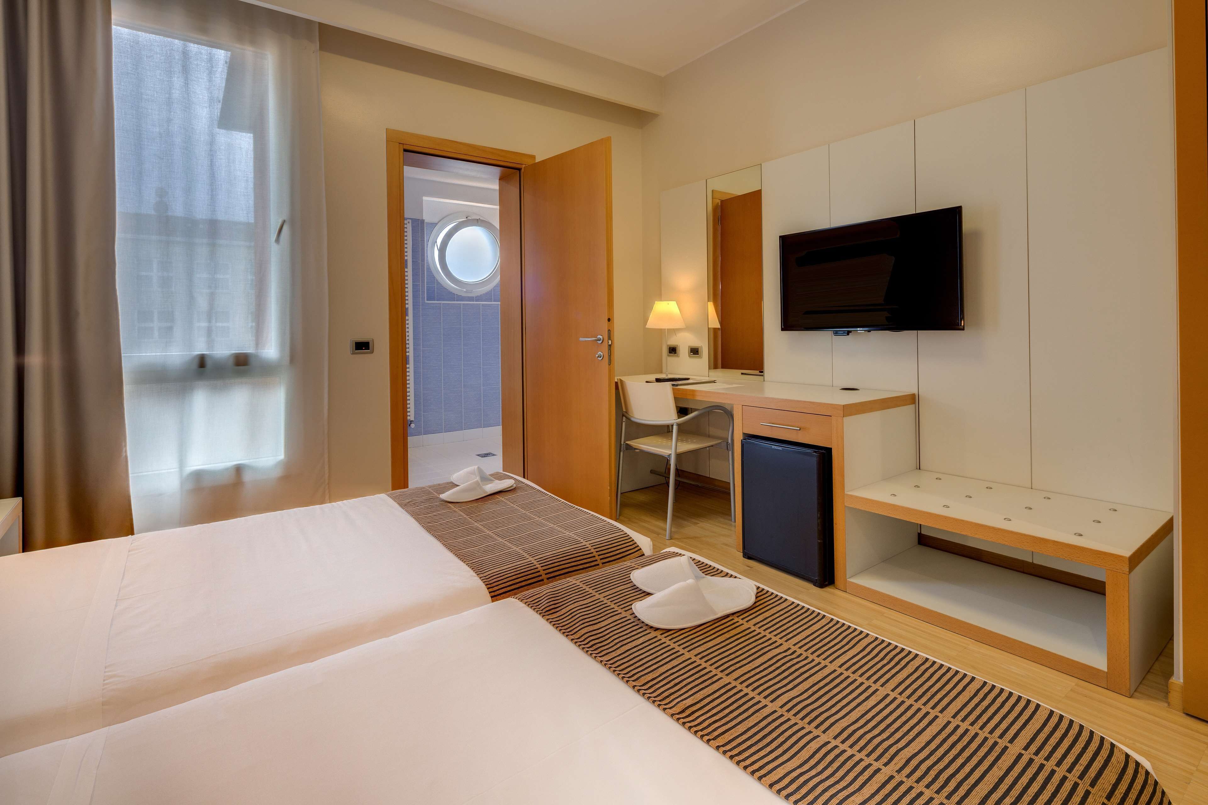 A hotel room with two beds, a desk, a television, and a bathroom with a window.