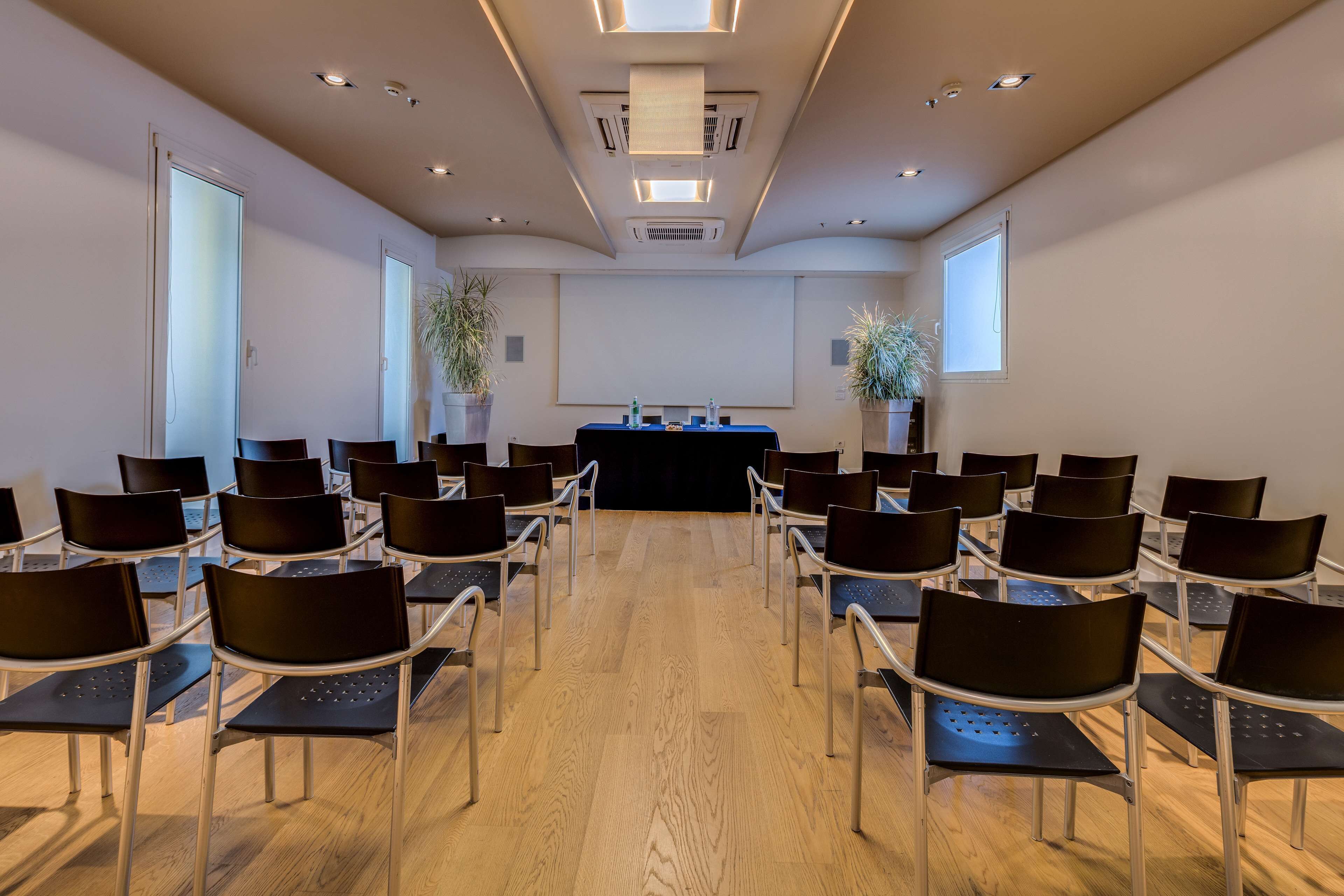 A conference room with rows of chairs facing a podium, wooden floors, and ceiling lights.