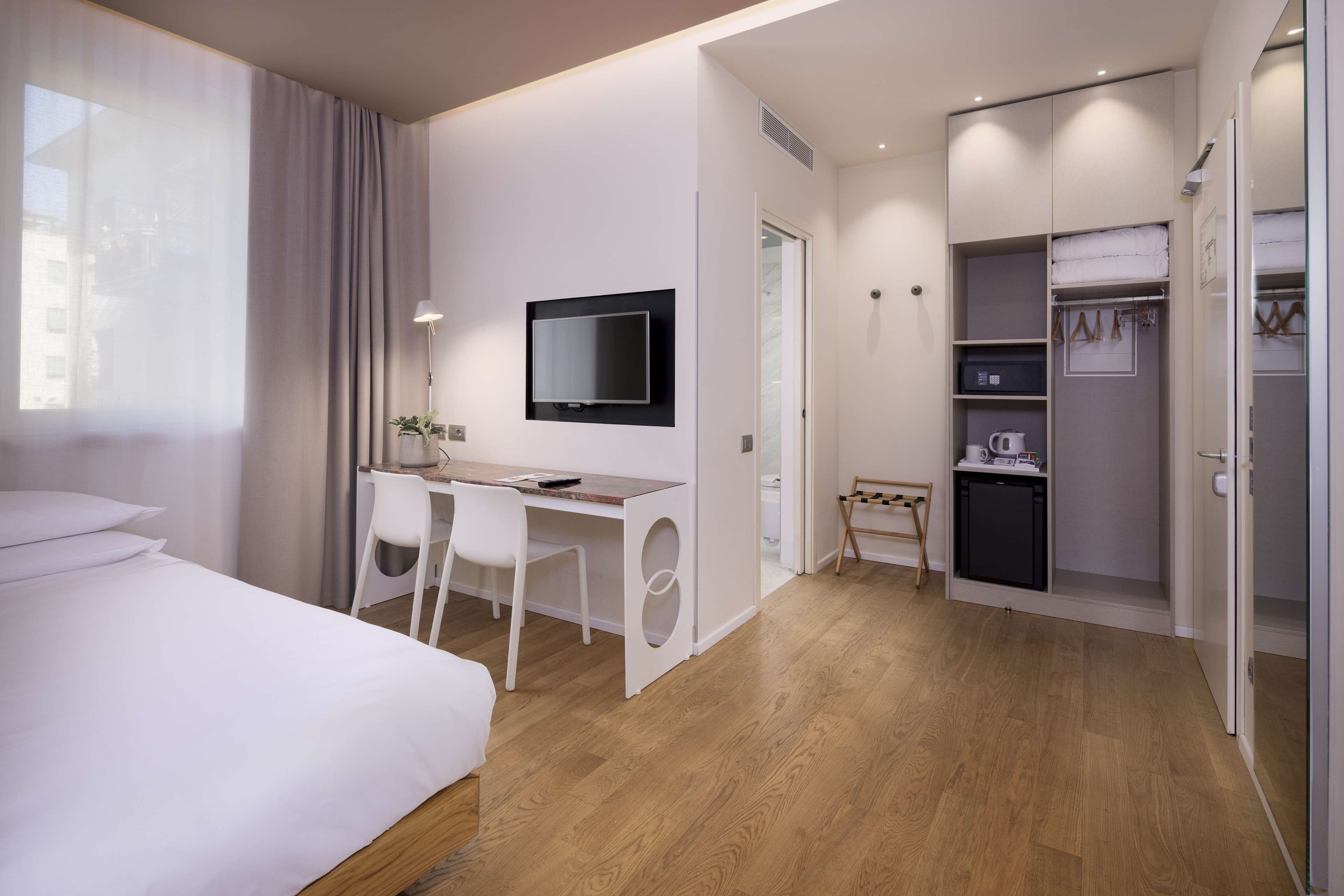 A modern hotel room with a bed, desk, chair, TV, and a walk-in closet with shelves and a drying rack.