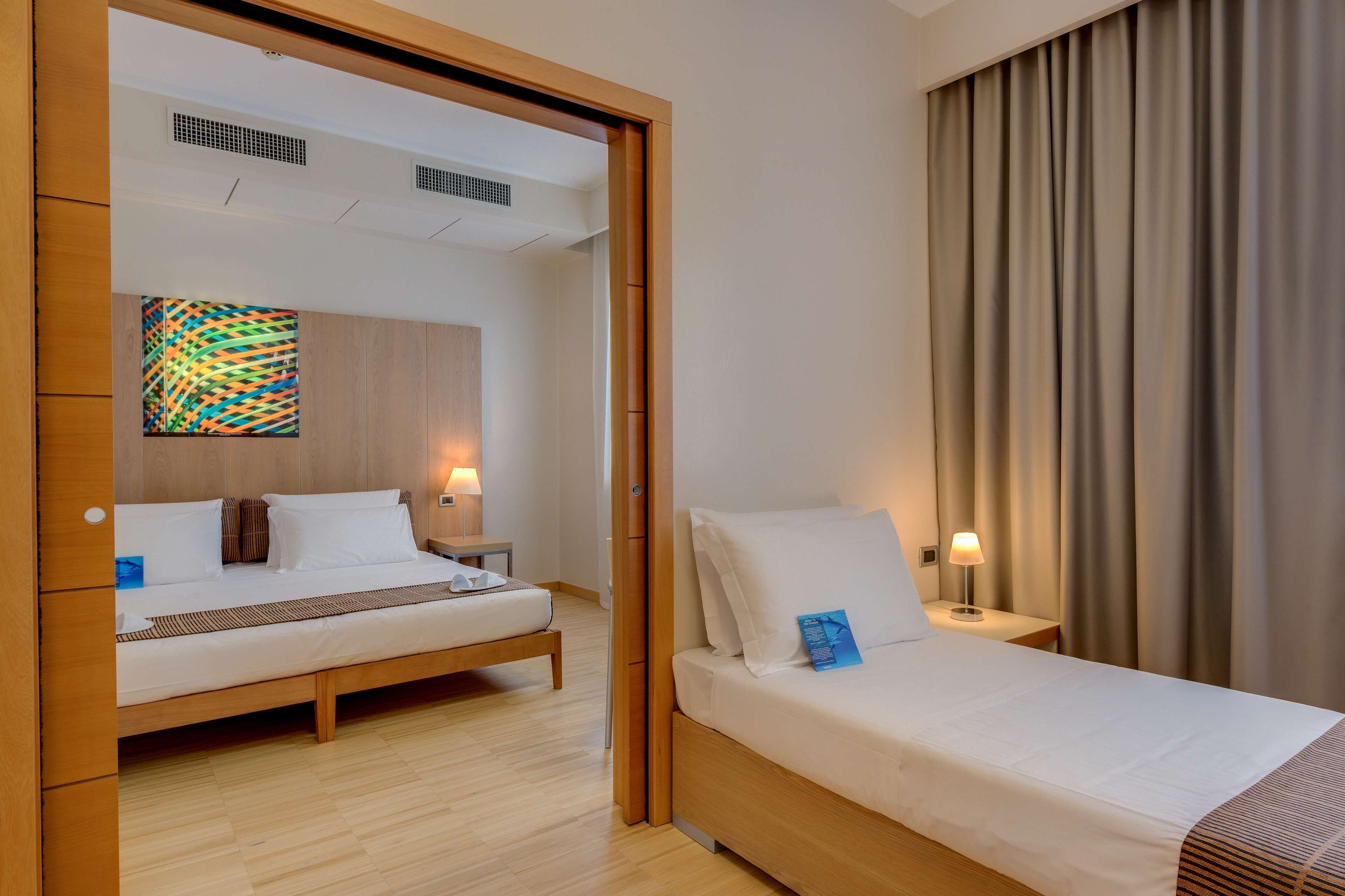A hotel room with two beds, wooden flooring, a large window with curtains, and a colorful abstract painting on the wall.