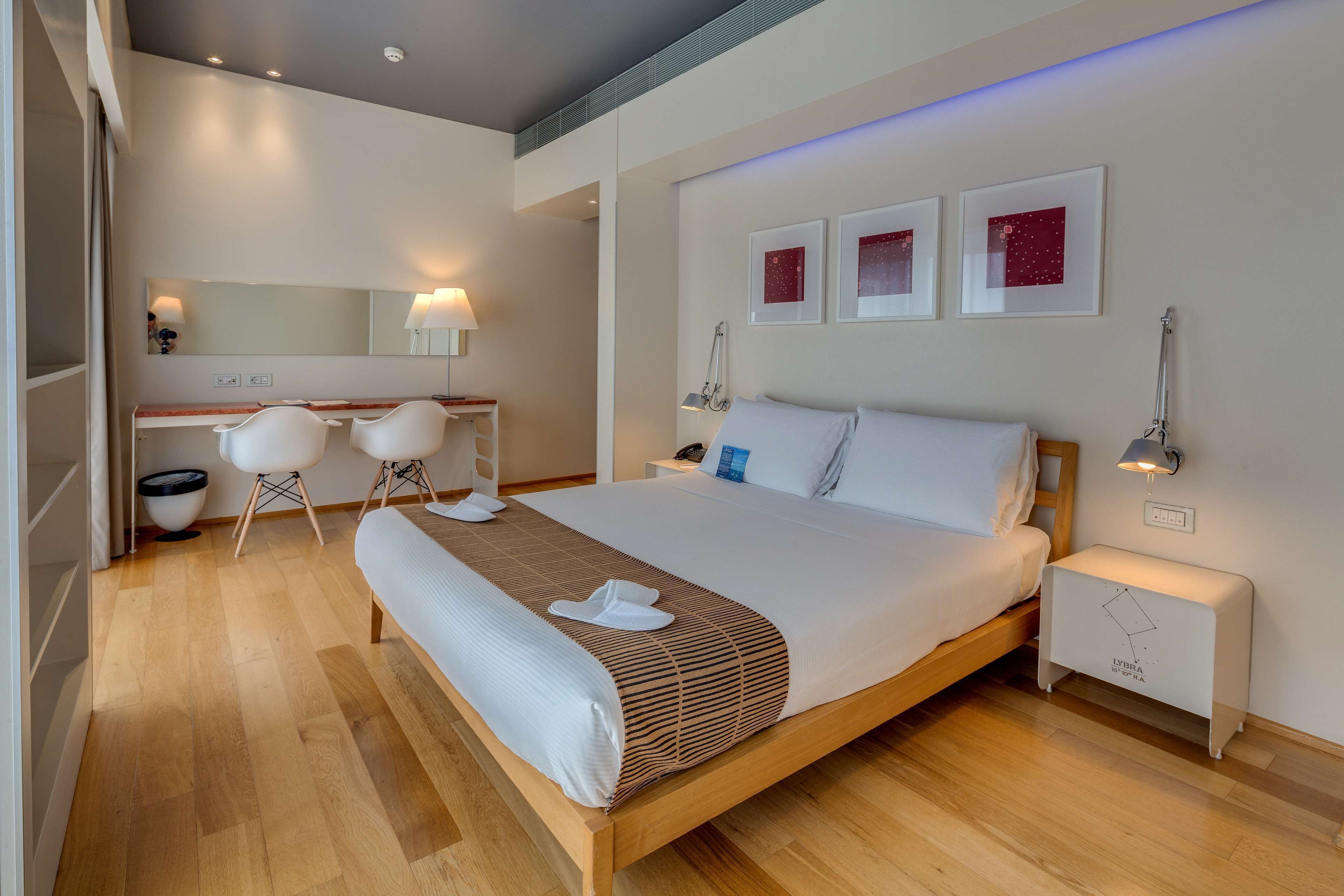 A modern hotel room with a wooden bed, a desk with chairs, a large mirror, and artwork on the wall.