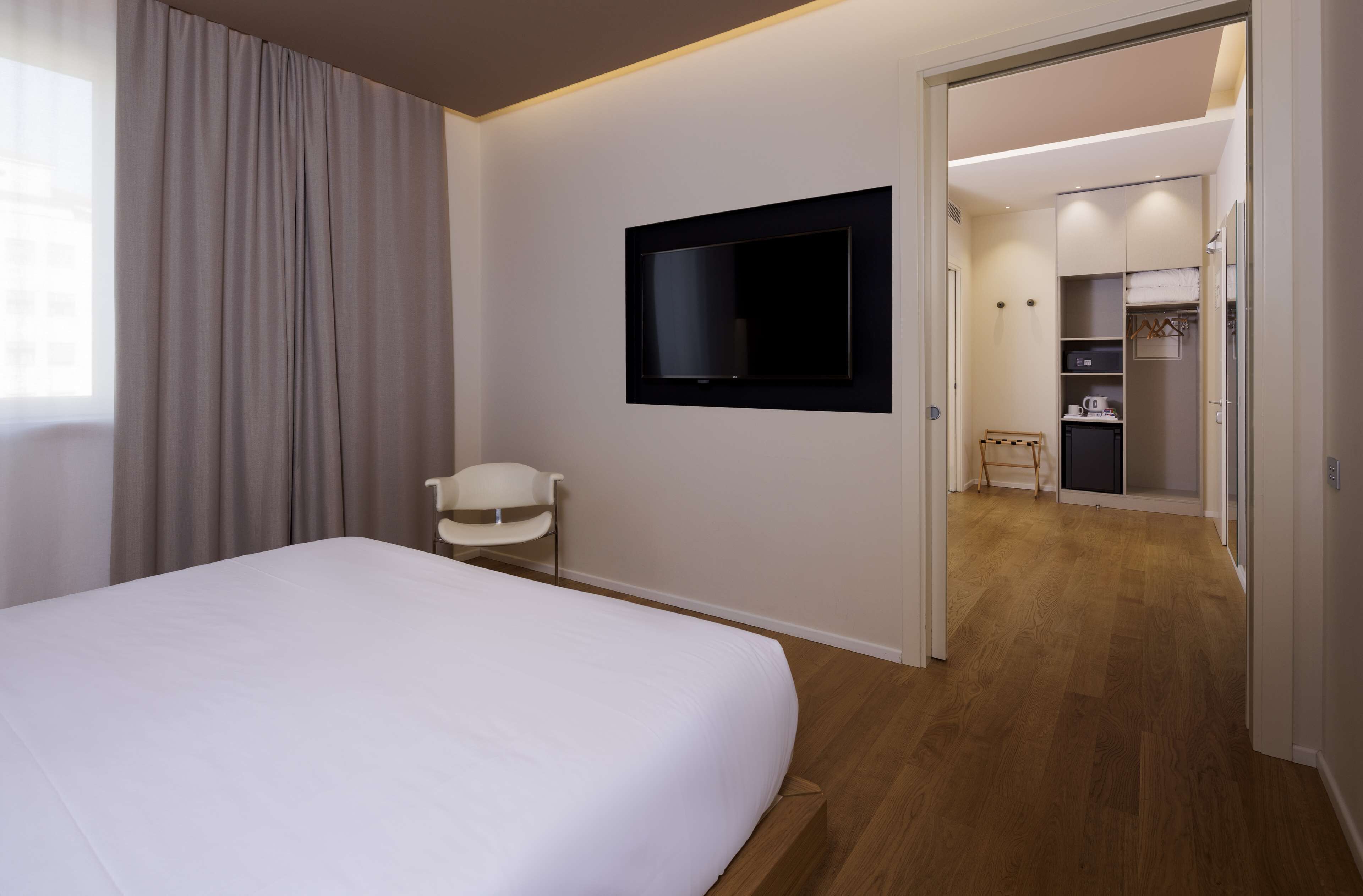 Habitación de hotel con cama, televisión montada en la pared, una silla y suelo de madera.