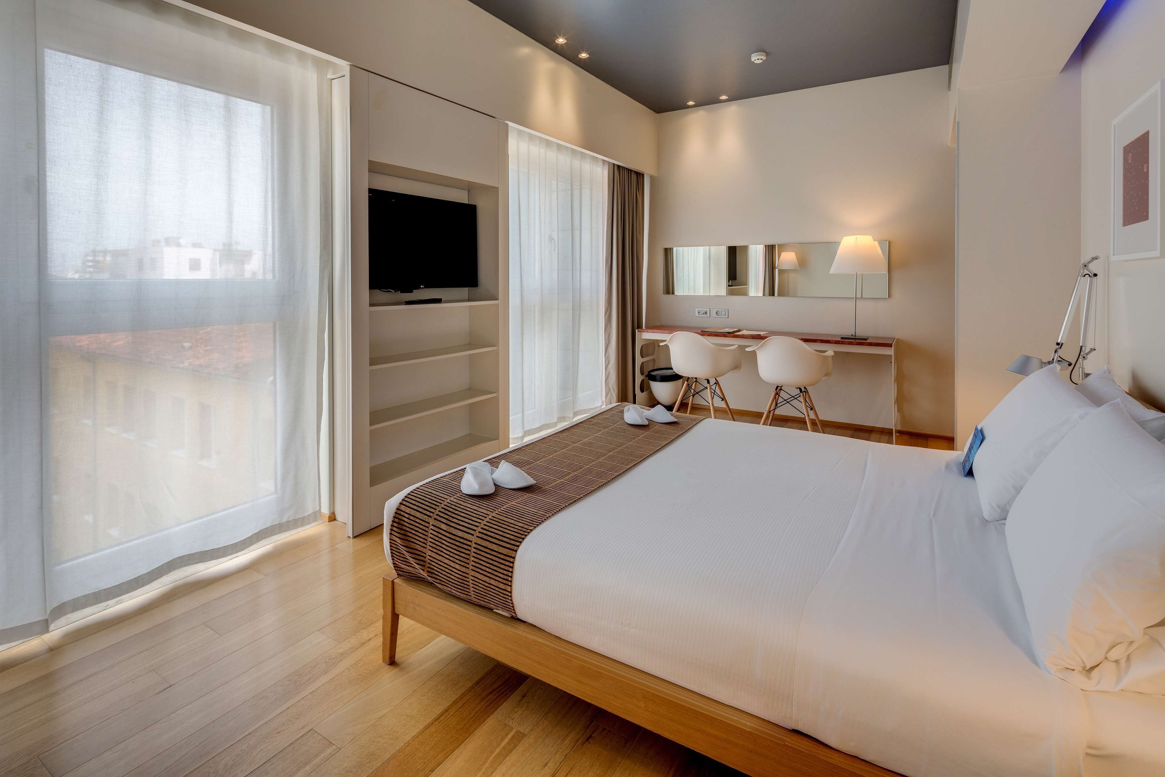 Ein modernes Hotelzimmer mit einem großen Bett, einem Schreibtisch mit Stuhl, einem Fernseher und einem großen Fenster mit Vorhängen.