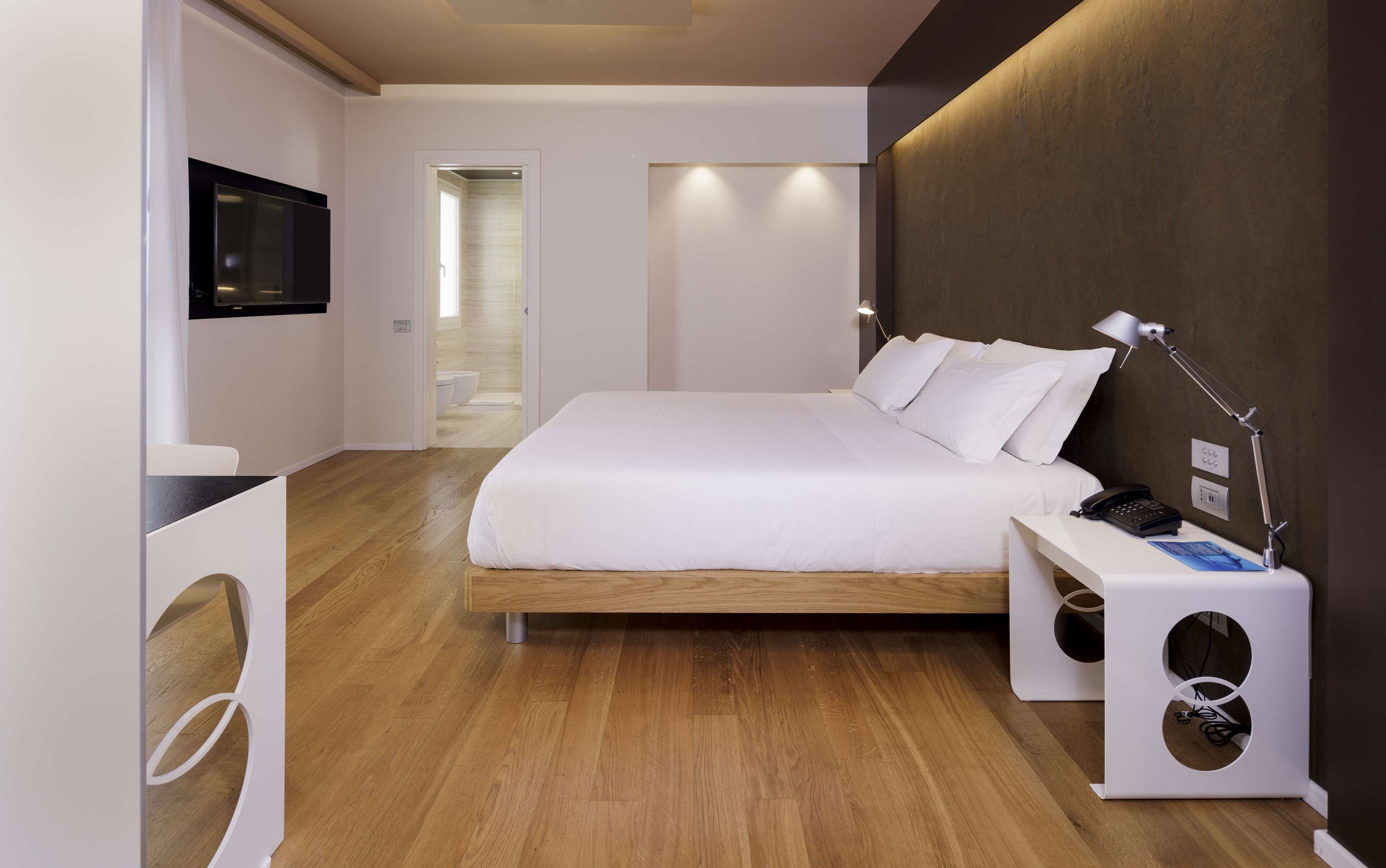 Quarto moderno com uma cama grande, uma mesa de cabeceira com abajur, TV fixada na parede e piso de madeira.