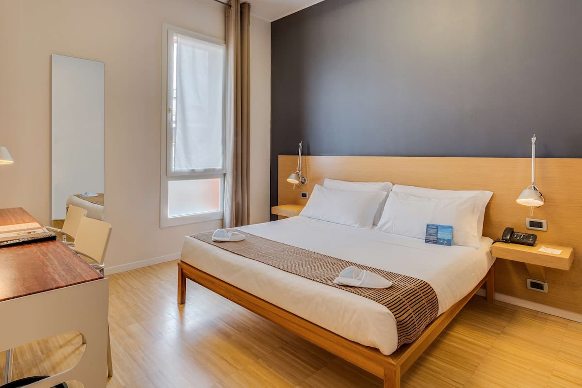 Una camera d'albergo con un letto di legno, comodini con lampade, una scrivania con sedia e un grande specchio.