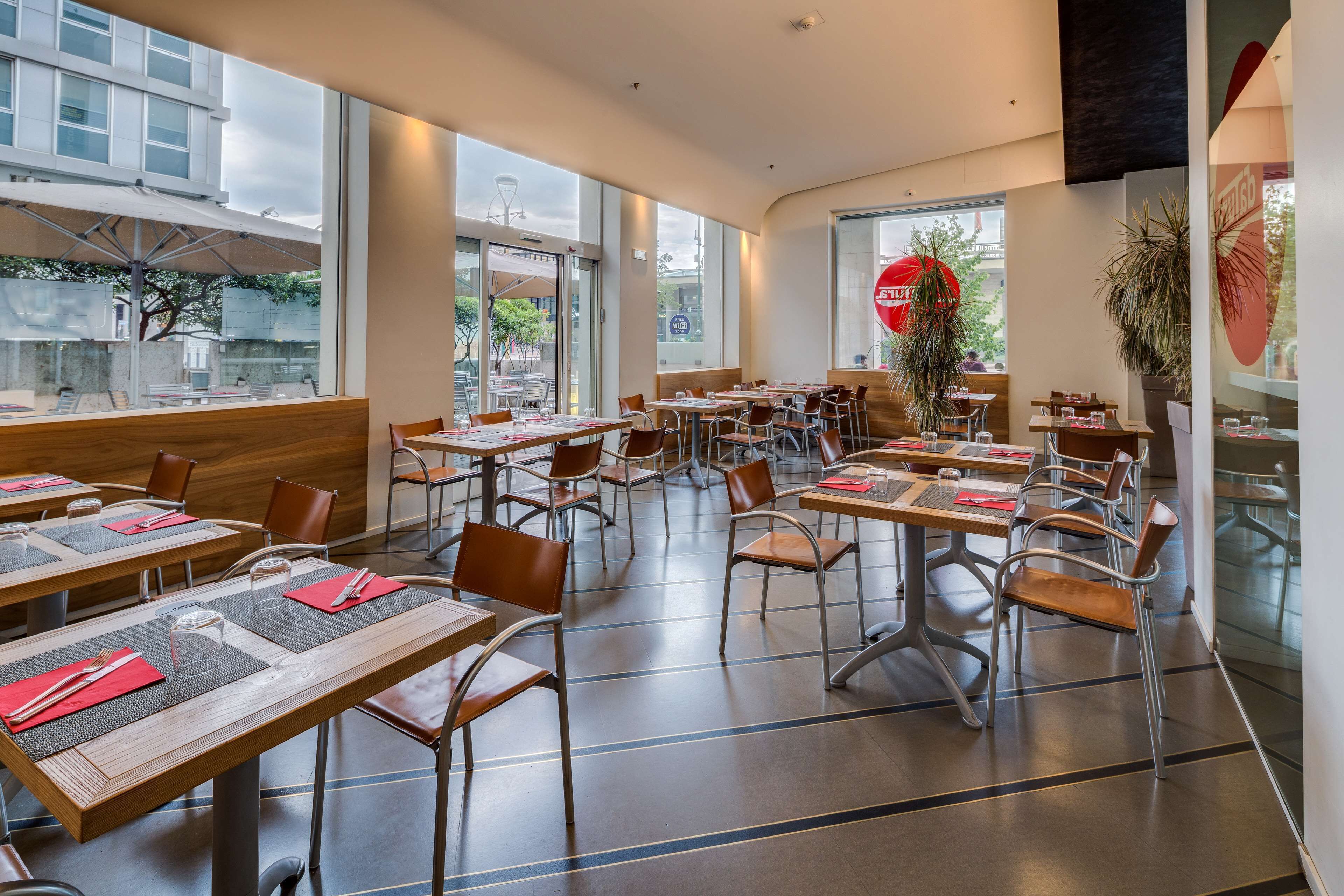 Un restaurante moderno con mesas altas, sillas, ventanas amplias y plantas de interior.