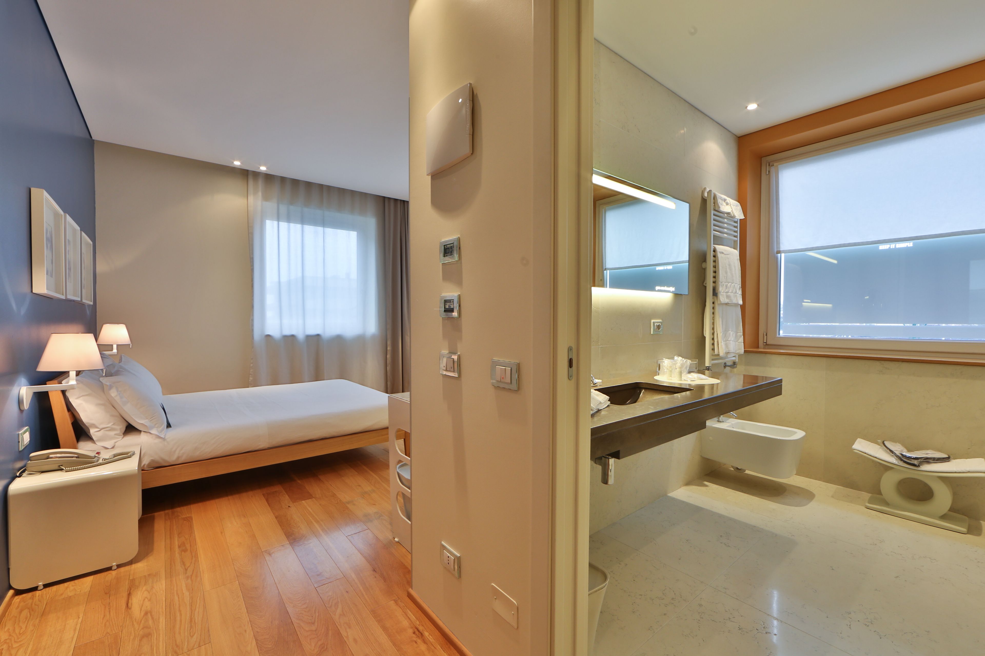 Quarto de hotel moderno com cama, mesa de cabeceira, abajur e banheiro com vaso sanitário e pia.