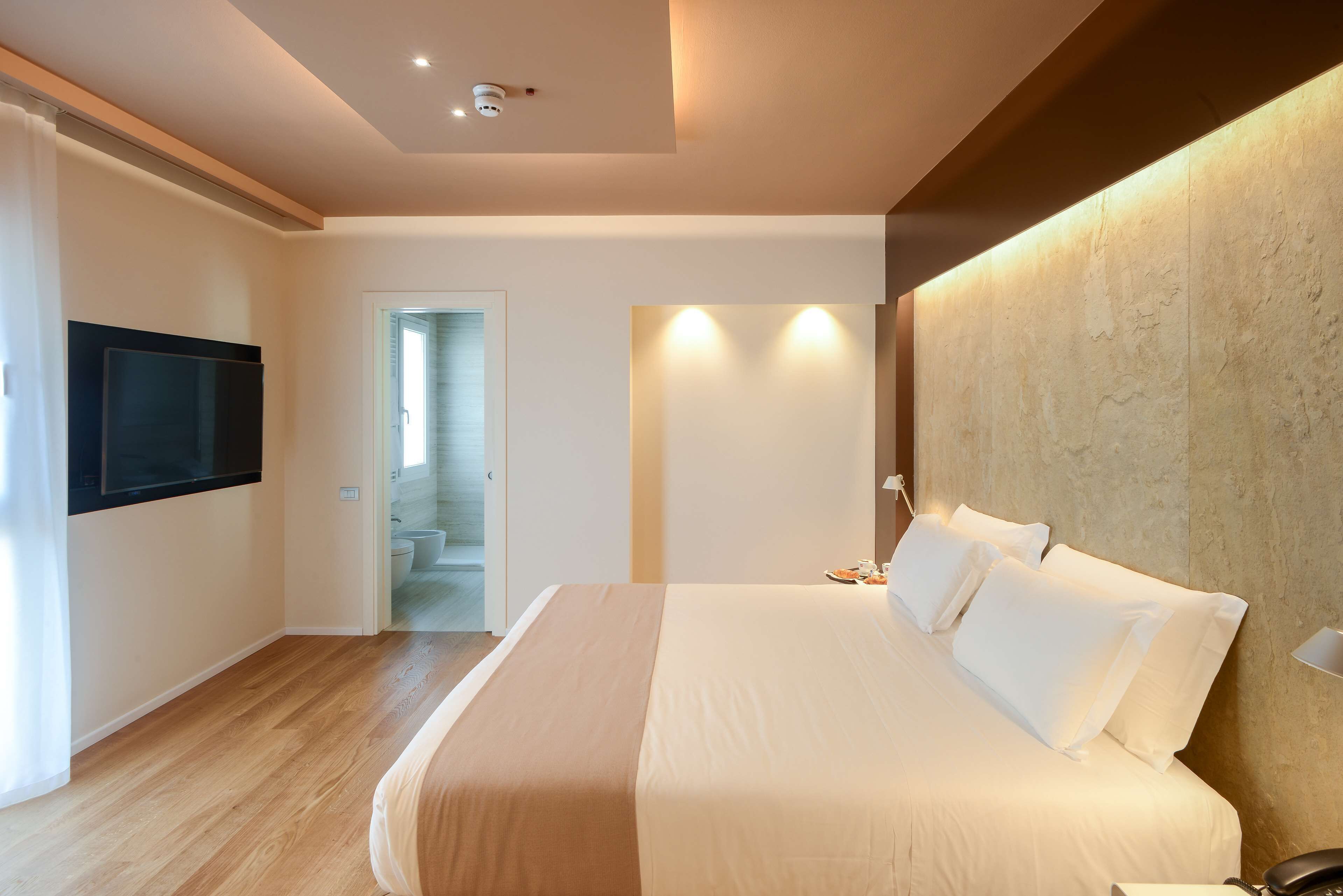 A modern hotel room with a large bed, a flat-screen TV mounted on the wall, and a wooden floor.