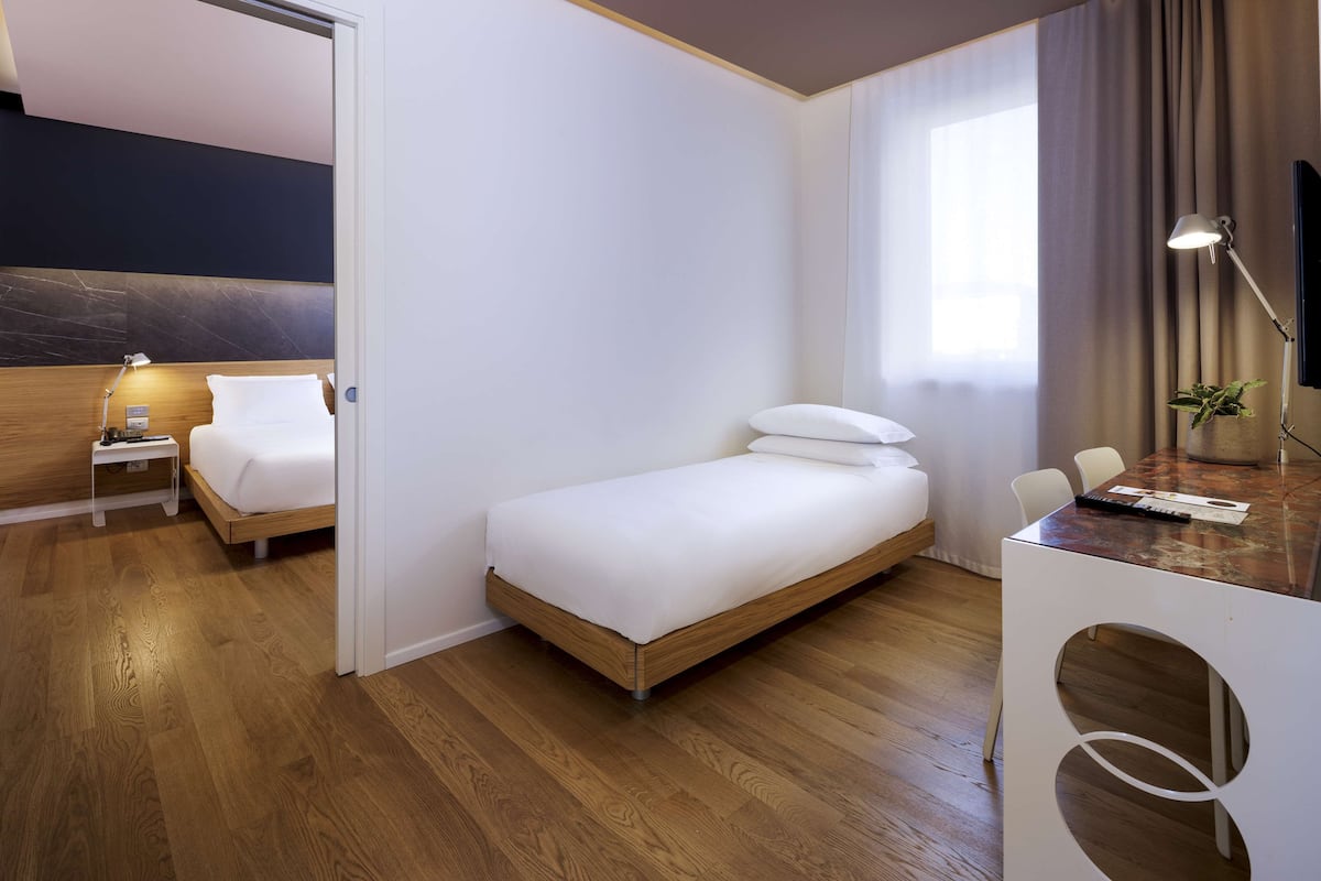 Una moderna camera d'albergo con pavimento in legno, un letto singolo, una scrivania con una sedia, un tavolino e un comodino con una lampada.