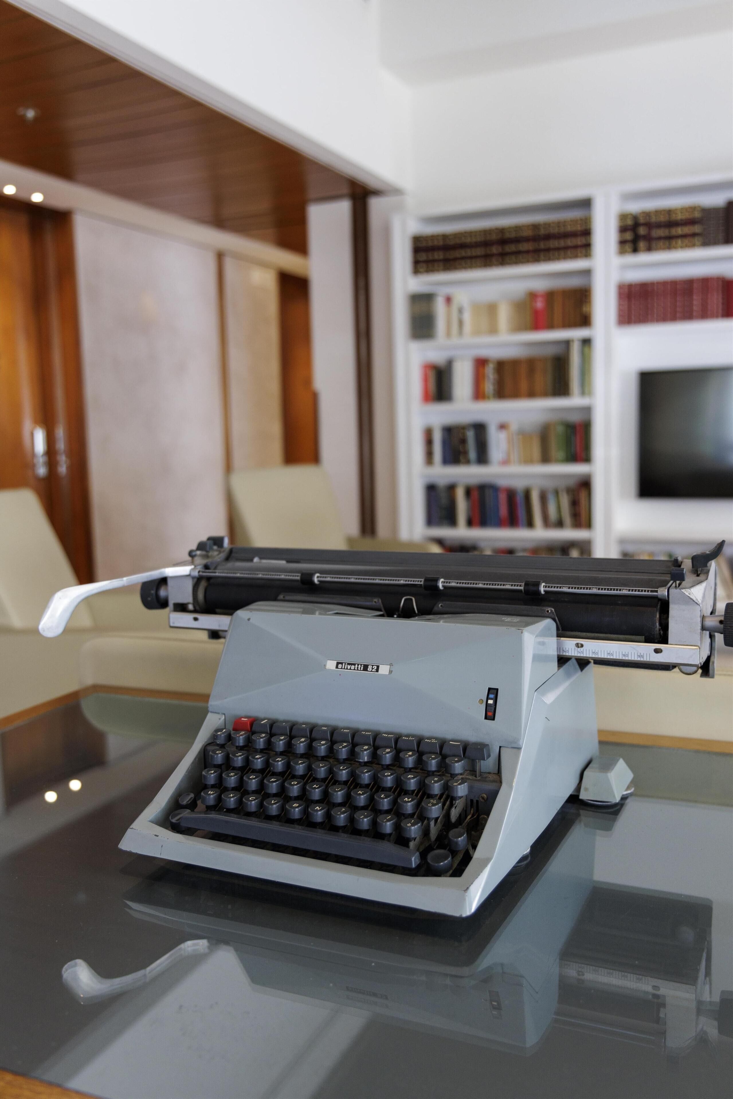 Une machine à écrire posée sur une table en verre, dans une pièce où l’on aperçoit une bibliothèque et un téléviseur au fond.