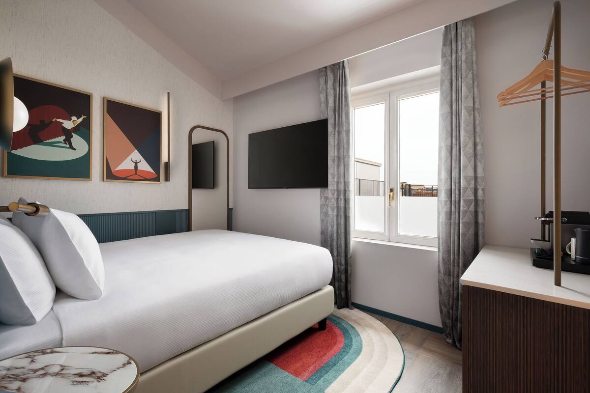 A modern hotel room with a bed, a flat-screen TV, a desk with a coffee machine, and a window with curtains.