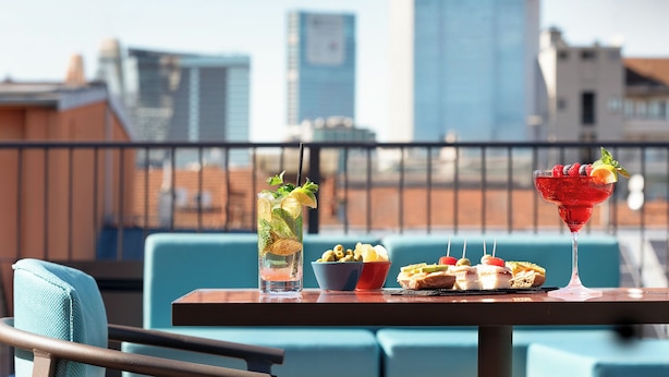 A rooftop bar with a city skyline view, a table with drinks and snacks, and blue chairs. - Puccini Hotel Milan, Tapestry Collection by Hilton (Milan)