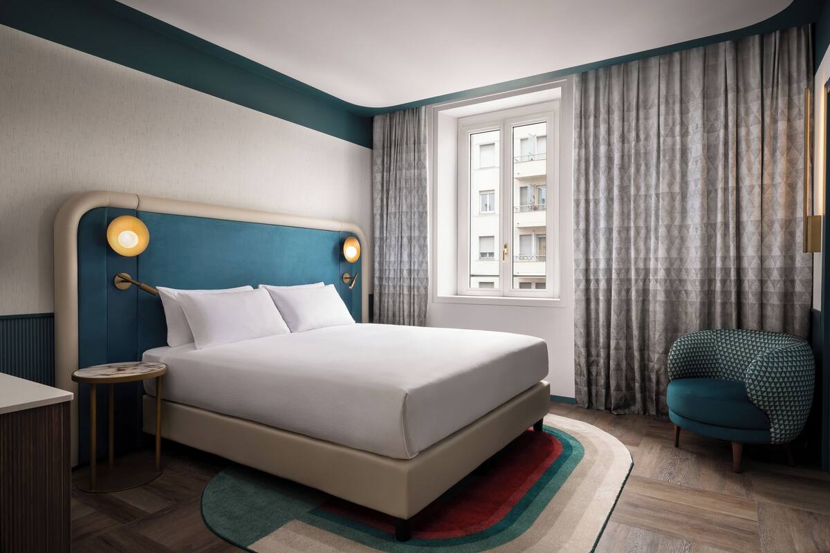 A modern hotel room with a large bed, a bedside table, a chair, and a window with curtains.