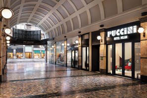A spacious corridor with a high ceiling and a tiled floor, leading to various retail stores including 'elfo puccini' and 'Hotel Milan'. - Puccini Hotel Milan, Tapestry Collection by Hilton (Milan)