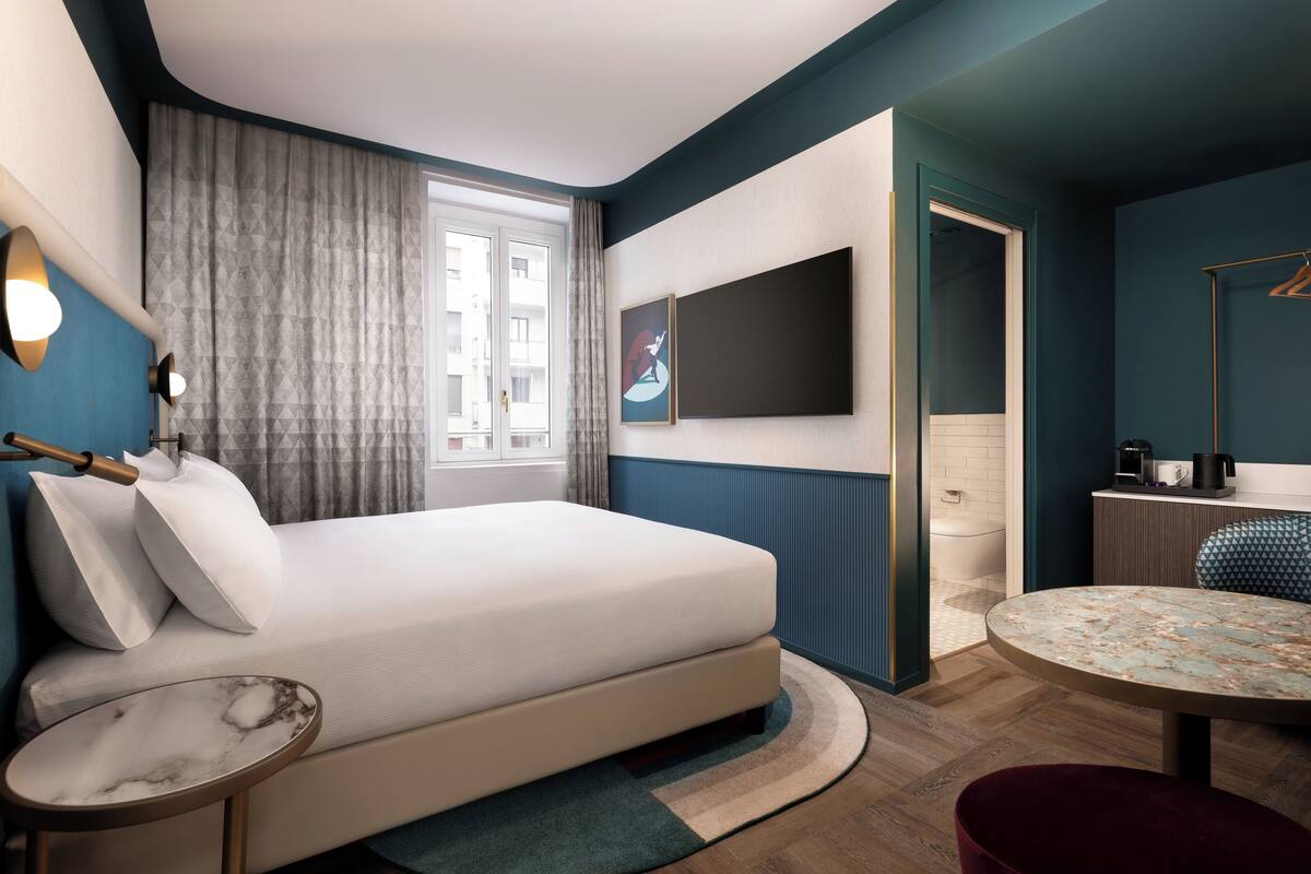 A modern hotel room with a bed, a round table, a small side table, a TV, and a bathroom visible through an open door.