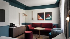 A modern hotel room with a red sofa, a round table, a small red ottoman, a hanging coat rack, and two abstract paintings on the wall. - Puccini Hotel Milan, Tapestry Collection by Hilton (Milan)