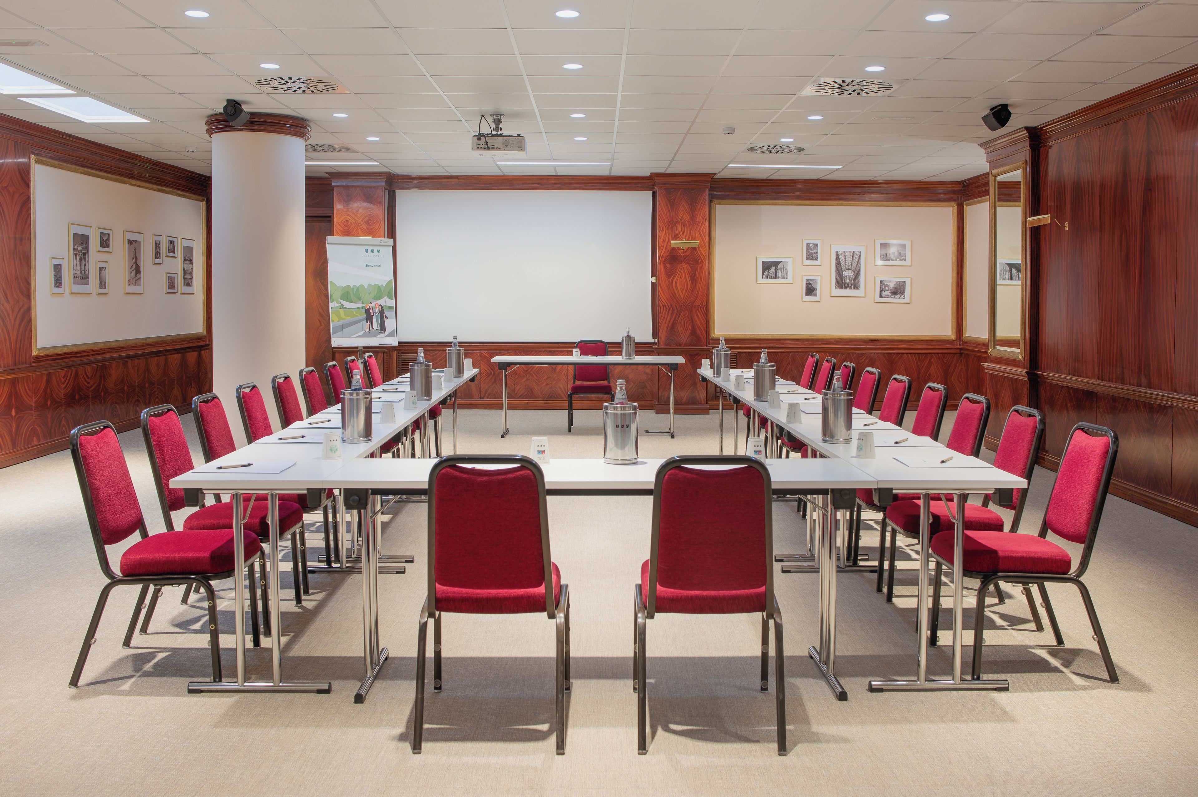 Un salón de conferencias con una larga mesa, sillas rojas, una pantalla de proyector y cuadros en las paredes.