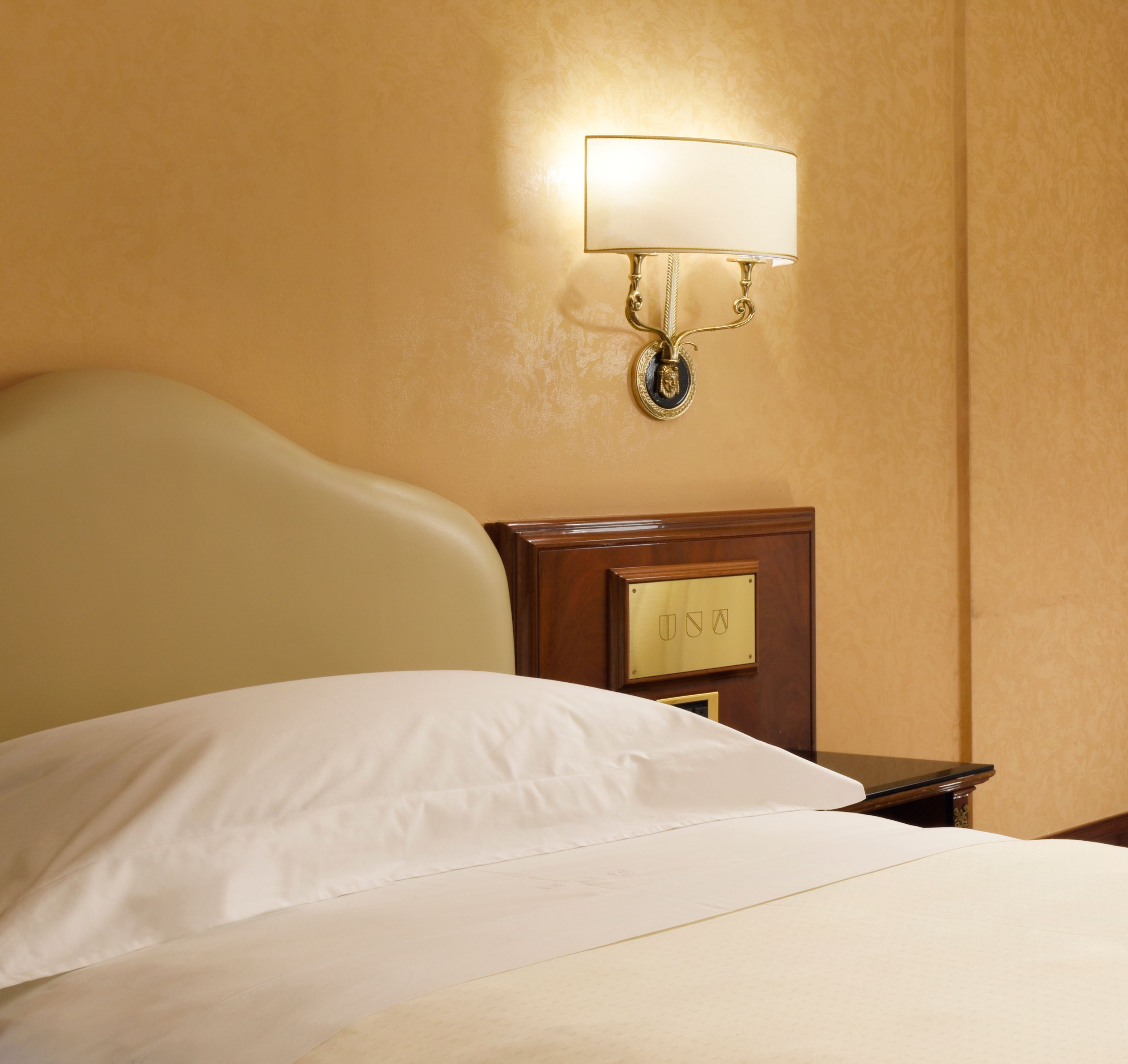 A hotel room with a bed, a nightstand, a wall-mounted lamp, and a wooden panel with a lock.
