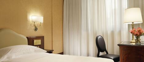 A hotel room with a bed, a desk with a lamp, a chair, and a nightstand with a lamp.
