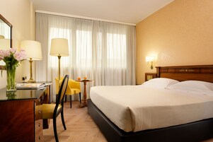 Een hotelkamer met een bed, een bureau met een lamp, een stoel, een kleine tafel met een lamp en een raam met gordijnen.