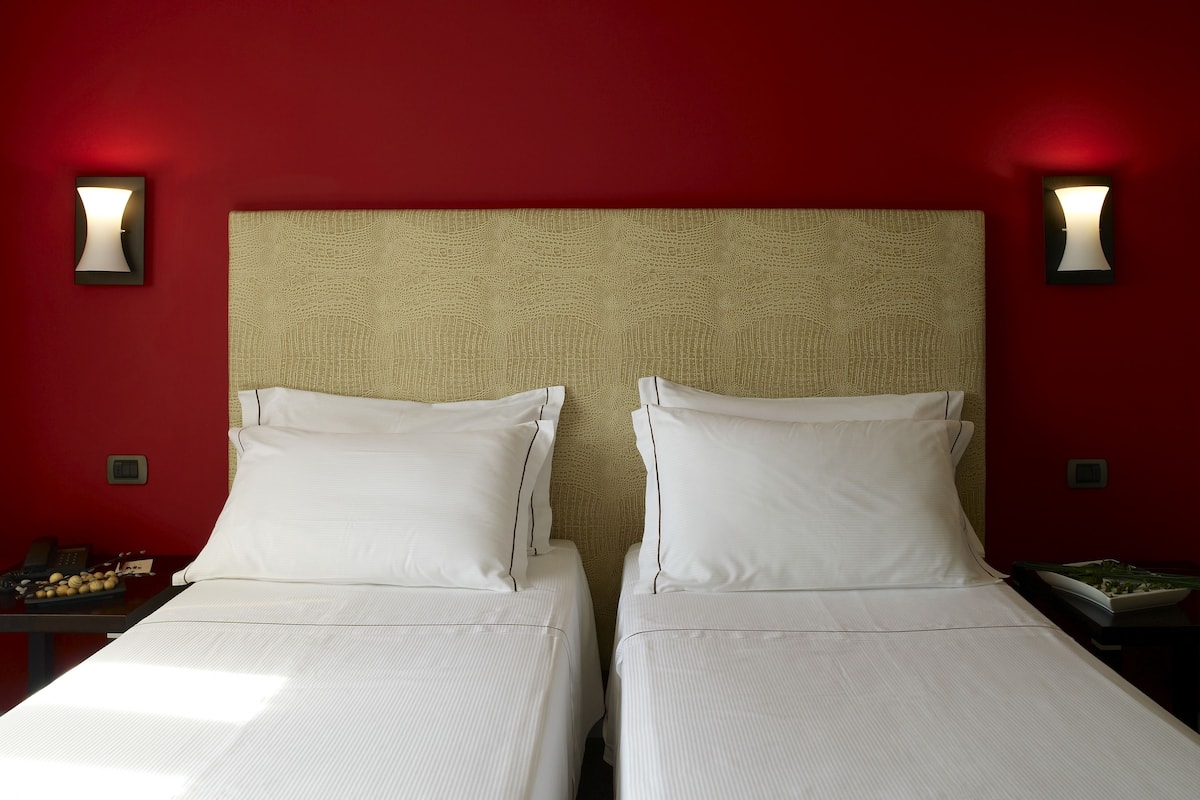 Habitación de hotel con dos camas, una pared roja y lámparas fijadas a la pared.