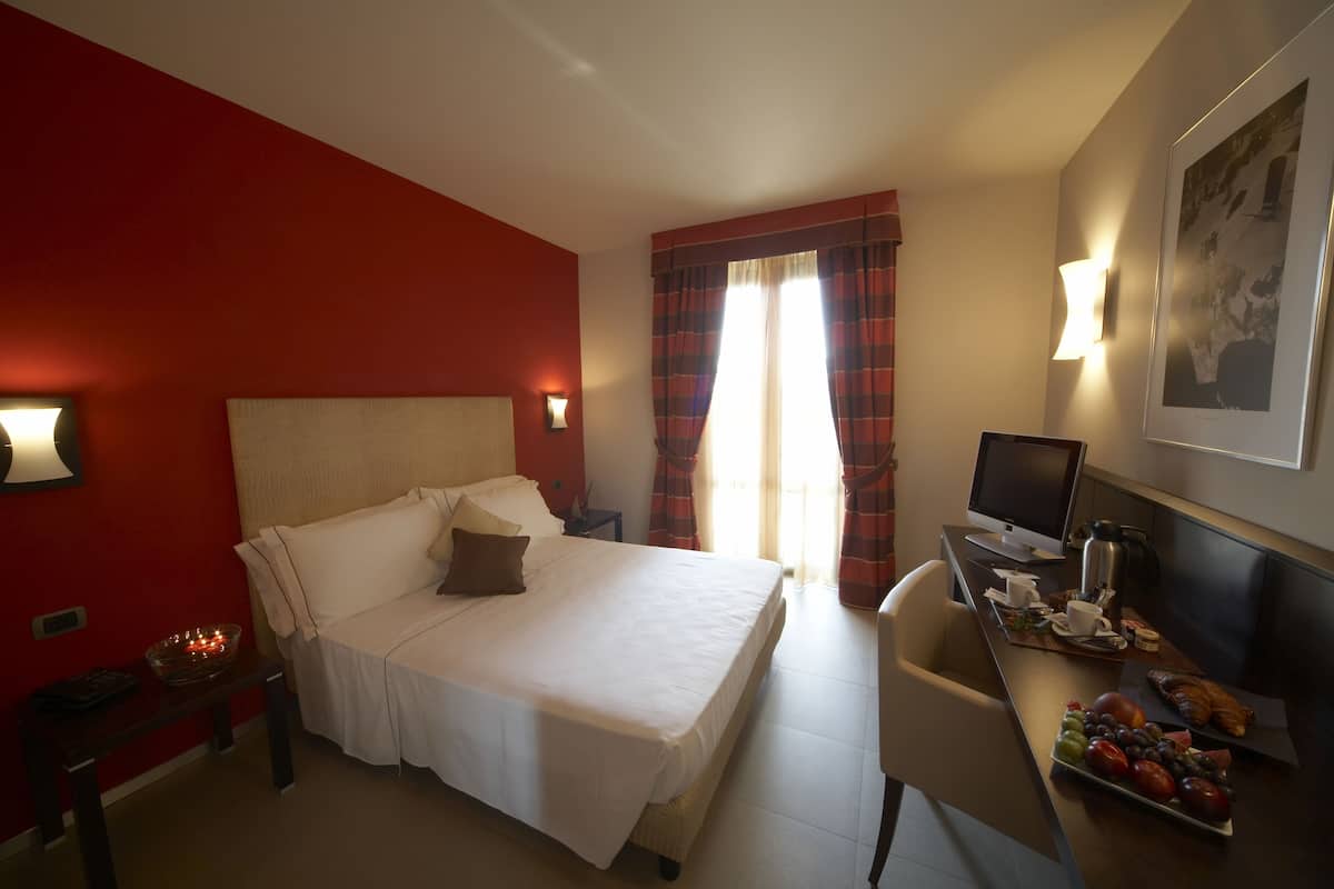 Habitación de hotel con una cama grande, un escritorio con computadora, una silla, una mesita con frutero, y una ventana con cortinas.