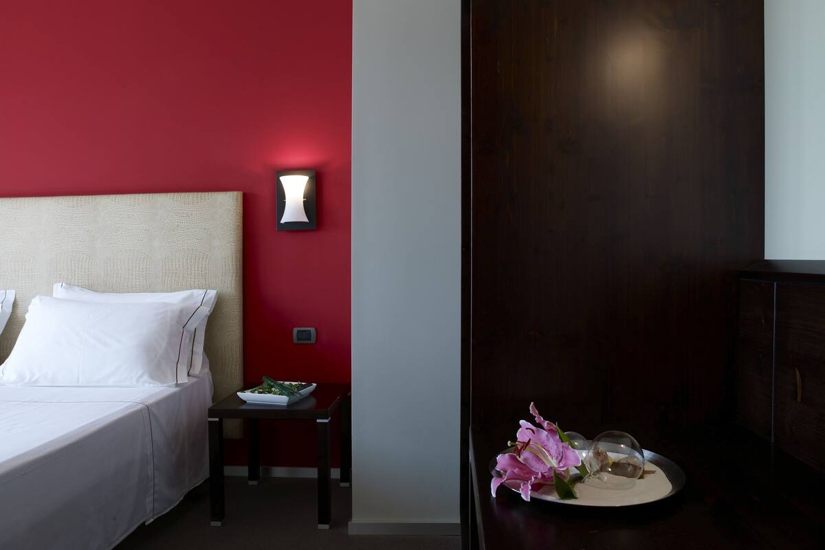 Habitación de hotel con una cama, una mesita con un frutero y una lámpara de pared, además de un adorno con flores.