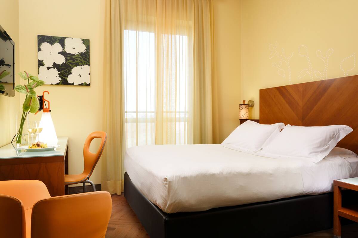 A hotel room with a bed, a desk with a chair, a potted plant, and a window with curtains.
