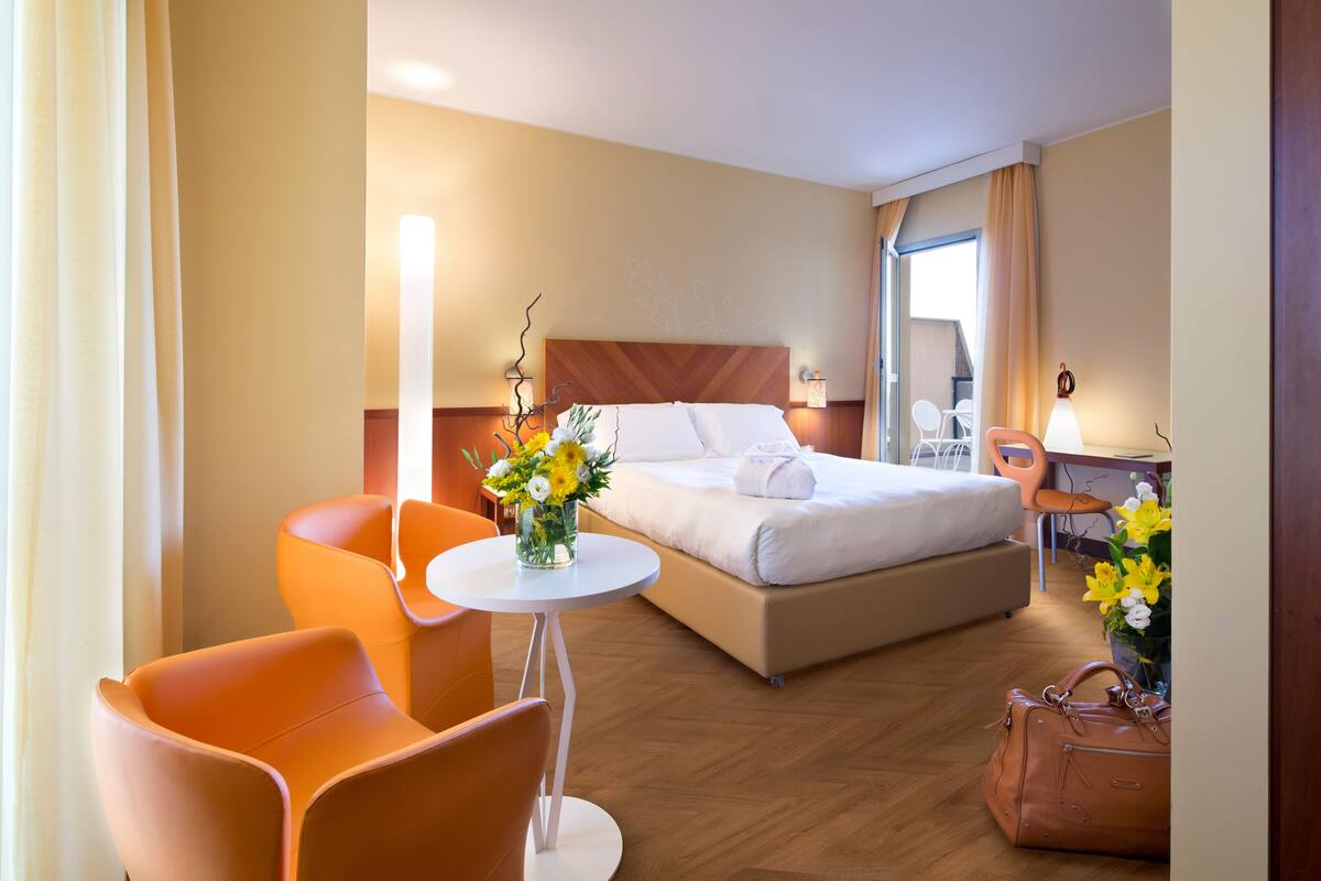A hotel room with a bed, two orange chairs, a small round table, and a suitcase.