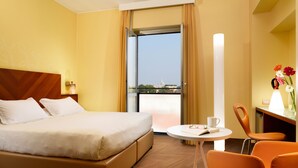A hotel room with a bed, a small table with a chair, a lamp, and a view of a cityscape through a window. - UNA Hotels Mediterraneo Milano (Milan)