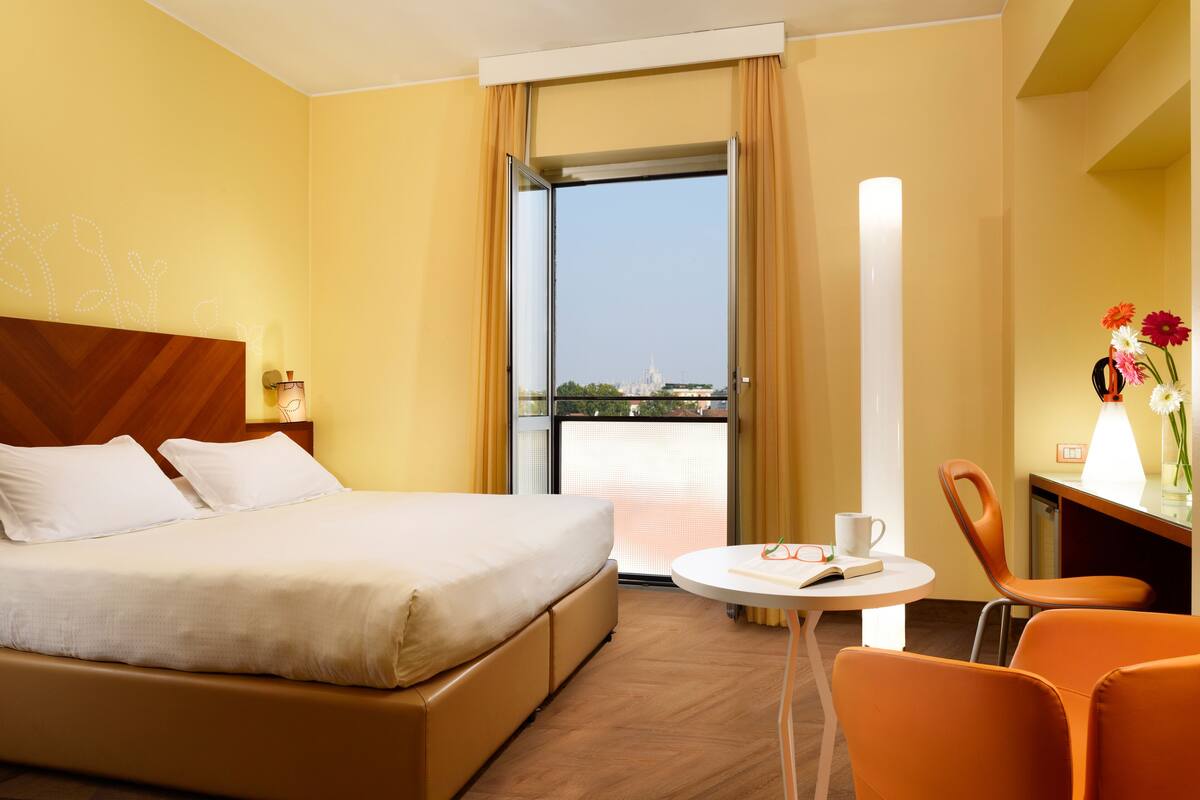 A hotel room with a bed, a small table with a chair, a lamp, and a view of a cityscape through a window.